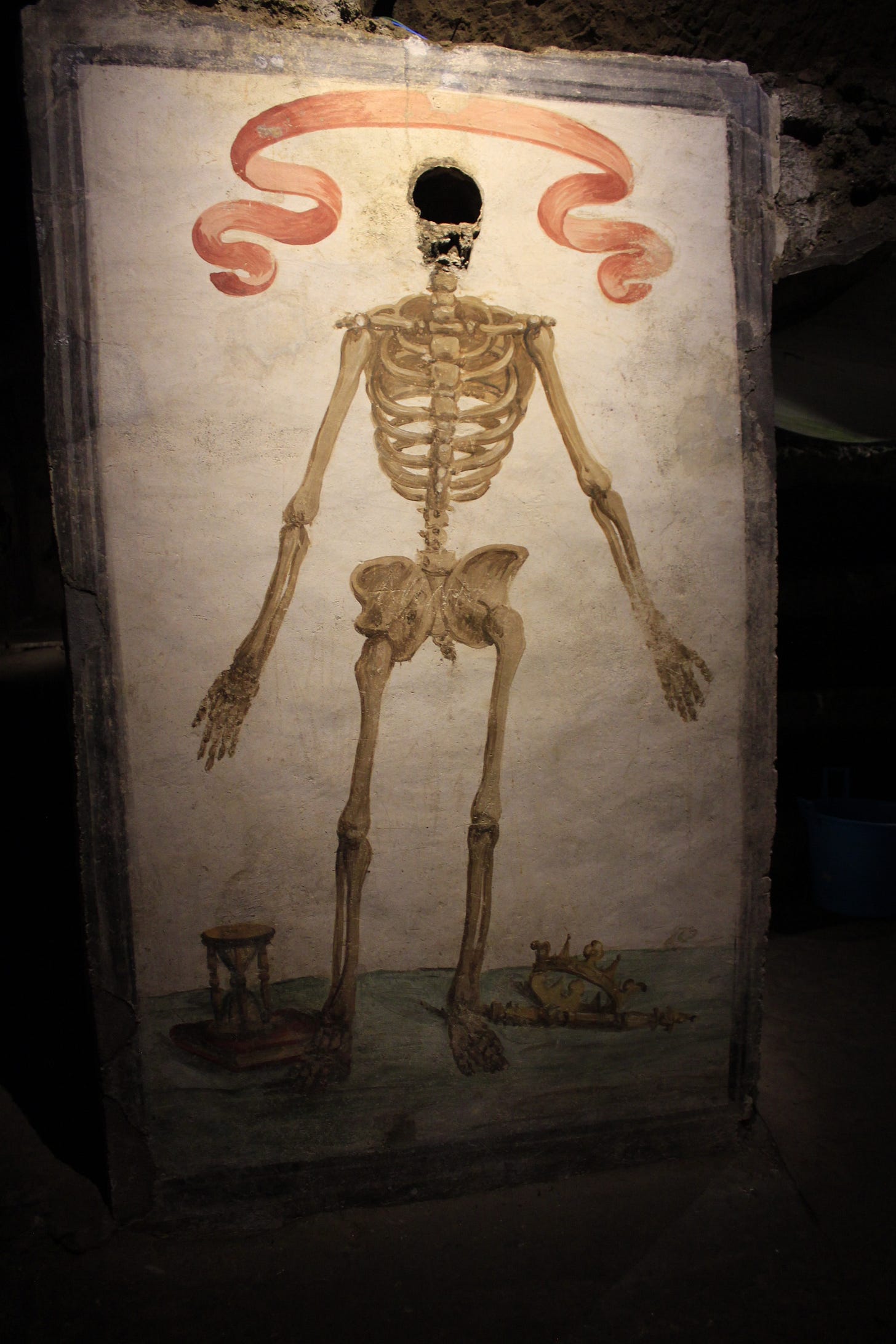 a fresco of a skeleton with a hole where a real skull was once mounted. there's a ribbon over its head and an hourglass and crown by its feet. a fresco of a skeleton with a hole where a real skull was once mounted. there's a ribbon over its head and an hourglass and crown by its feet.