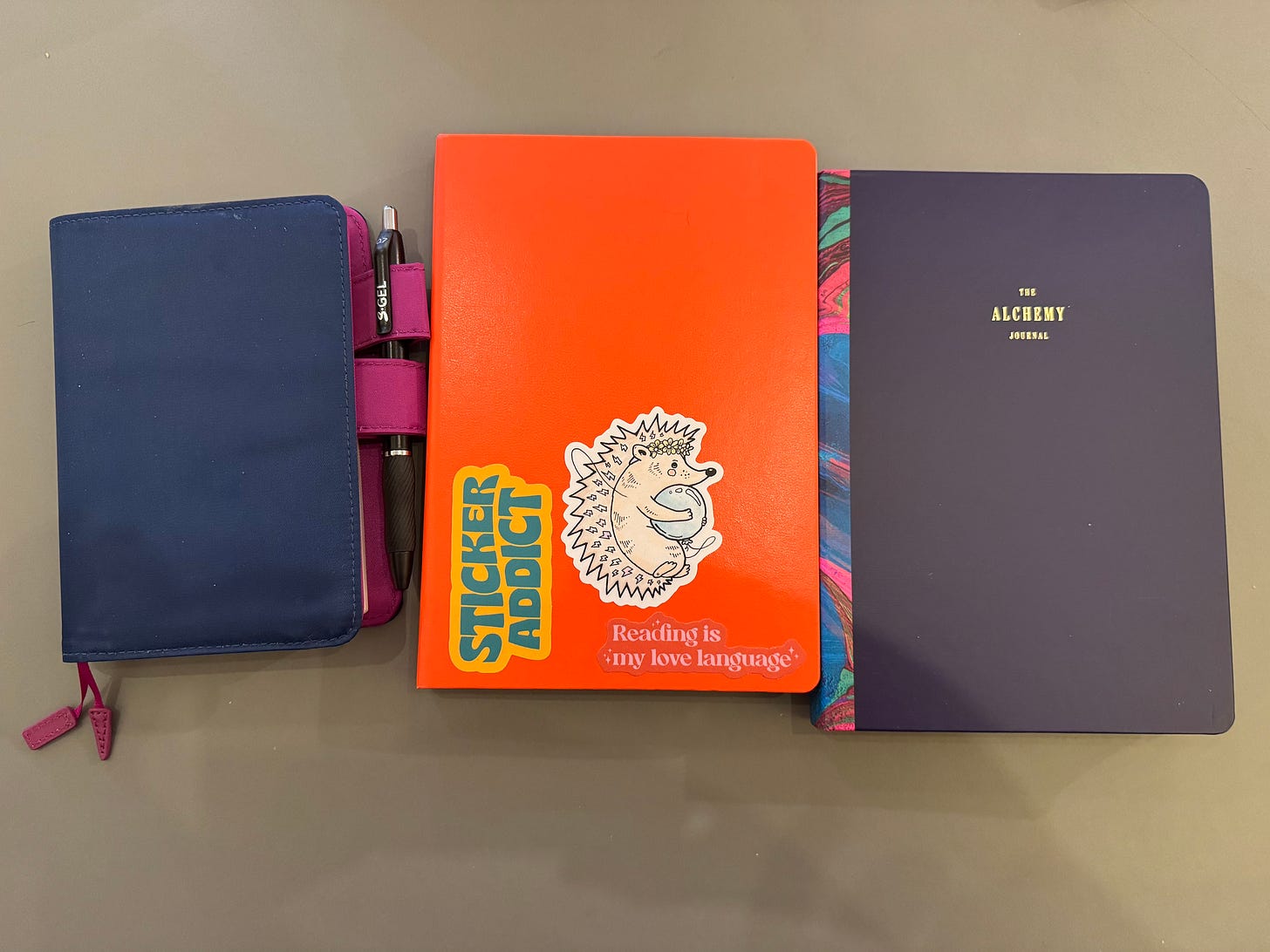 Three notebooks on a greenish table - A6 dark blue with pink accents, bright orange A5, and dark blue A5