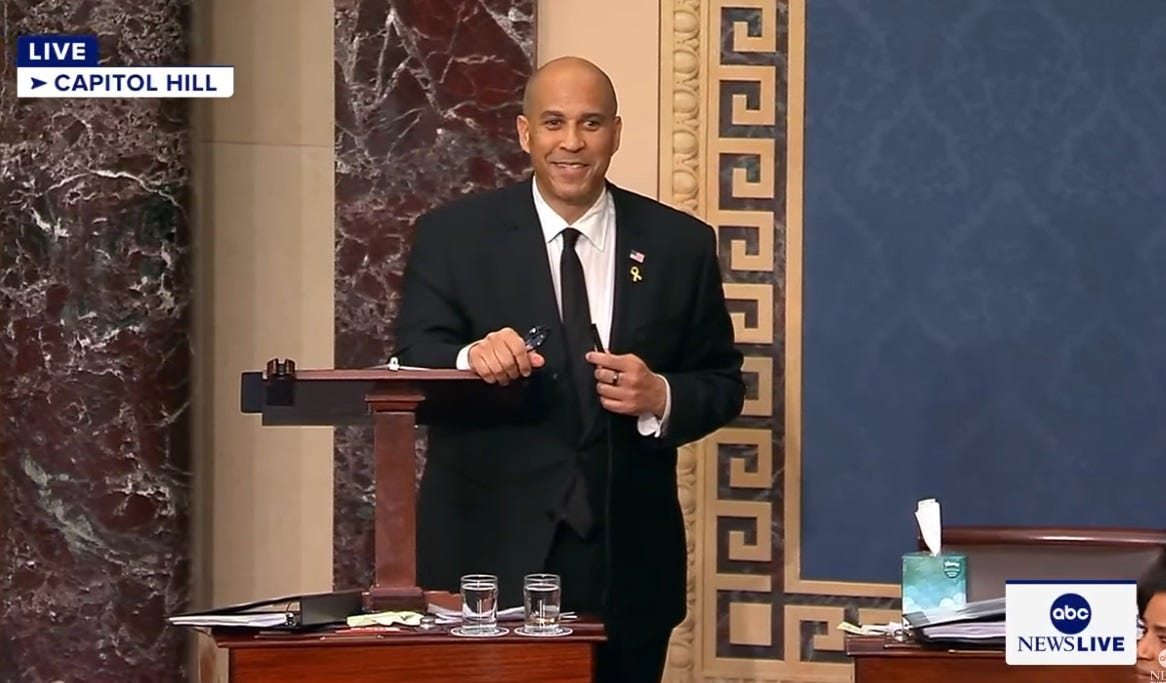 Sen Cory Booker smiles as he prepares to yield the floor, standing at the lectern in the Senate and holding the clip-on microphone he's just removed for the first time in 25 hours