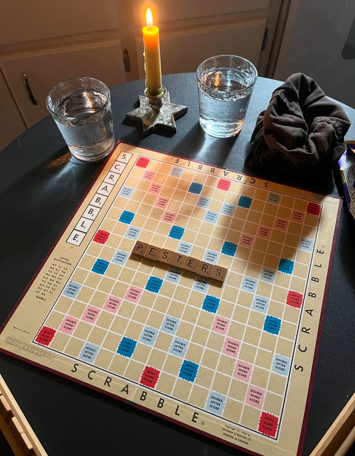 A little table with a scrabble game on it. There's just one word on the board, and it is "PESTERS". Also on the table are a couple glasses of water and a lit candle.