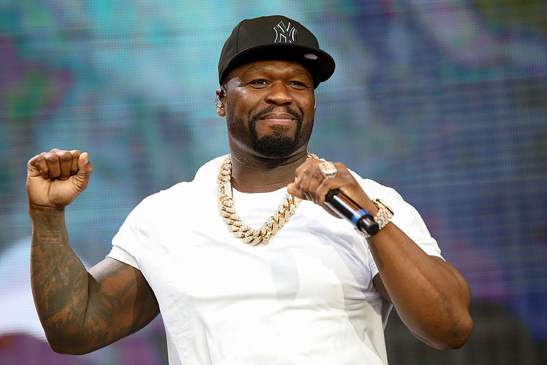 50 Cent performing on stage wearing a white t-shirt, black baseball cap, and gold chain, holding a microphone with both fists raised