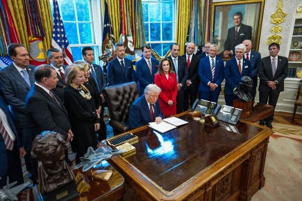 President Trump sits at his desk in the Oval Office, surrounded by lawmakers and automobile executives.