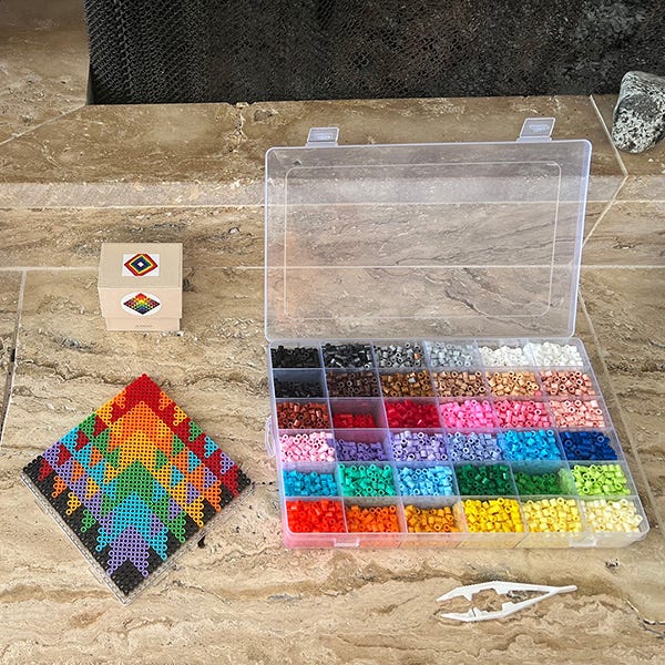 A photo of an art kit filled with plastic beads that melt when you iron them together called fuse beads. This is sitting on a fireplace along with a composition of fuse beads laid out like the rainbow squared icon.