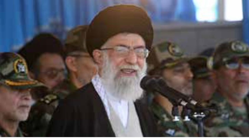 Color photograph of Iran’s Supreme Leader Ali Khamenei, with a white beard and glasses, wearing a dark robe and turban, speaking into a microphone. He stands in front of several uniformed military personnel in camouflage, who are positioned closely behind him in what appears to be a formal or official setting.