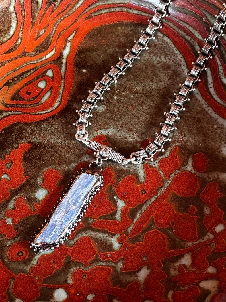 May include: A silver chain necklace with a blue crystal pendant. The pendant is set in a silver bezel and has a textured surface. The necklace is on a red and brown patterned surface.