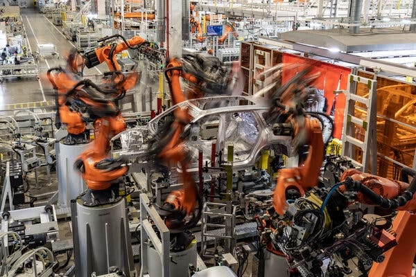 Orange robotic arms, blurry because they are in motion, surround the body of a car in a factory. 