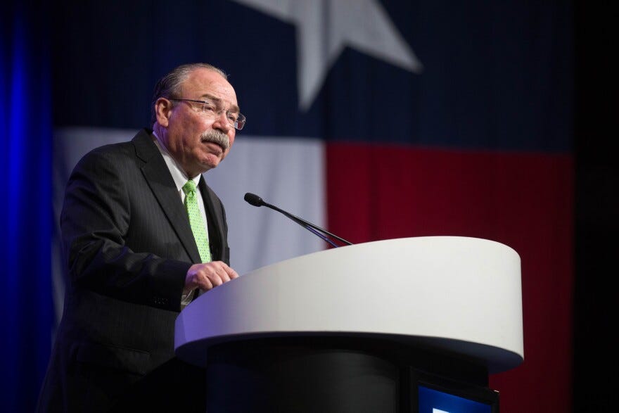 Texas Democratic Party Chairman Gilberto Hinojosa speaks behind a podium with a Texas flag behind him at the 2018 Texas Democratic Convention in Fort Worth, Texas. He is wearing a suit with green tie and eyeglasses.