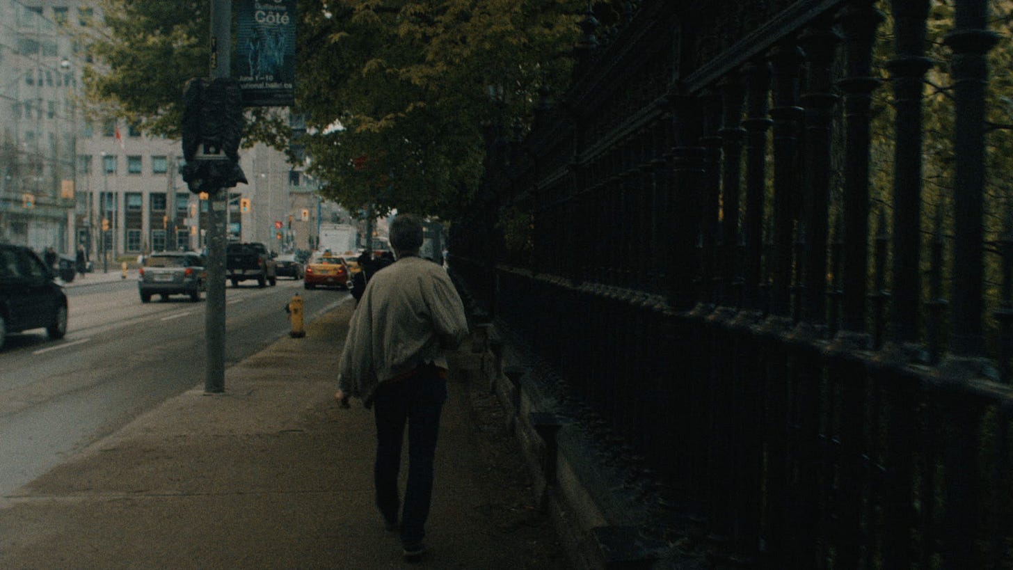 Person walking along a dim city sidewalk beside a black iron fence.