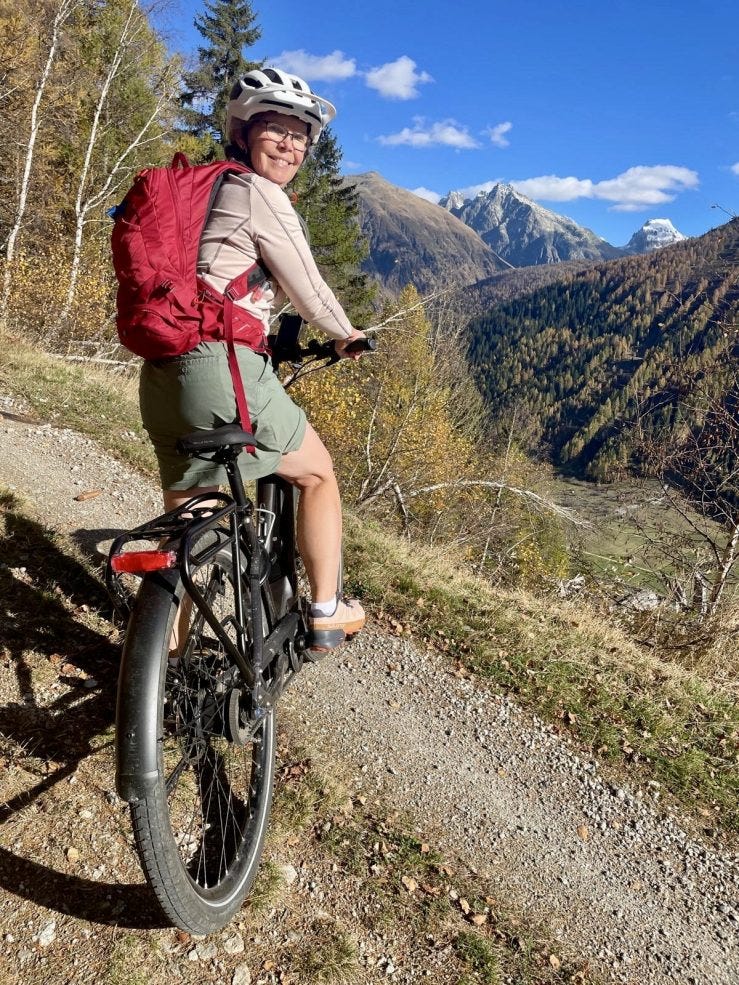 A woman straddles an city e-bike on a mountain gravel road. She's in a green tech-fabric shirt and a long-sleeved pink tech shirt. Her backpack is red and her helmet's white. She wears glasses and her hair's gray. She's looking over her should to smile at the camera. In the background is the valley floor of Obergoms and snow-capped peaks. The larch and birch needles and leaves are yellow. It's fall in the Alps.