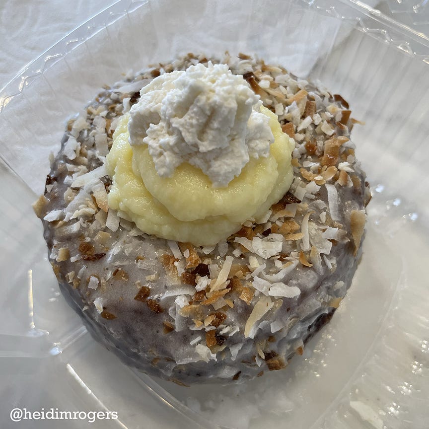 Close-up of a decadent donut, glazed and coated in toasted shredded coconut with coconut cream pie filling and whipped cream on top. It sits in a clear plastic tray on a white tabletop.