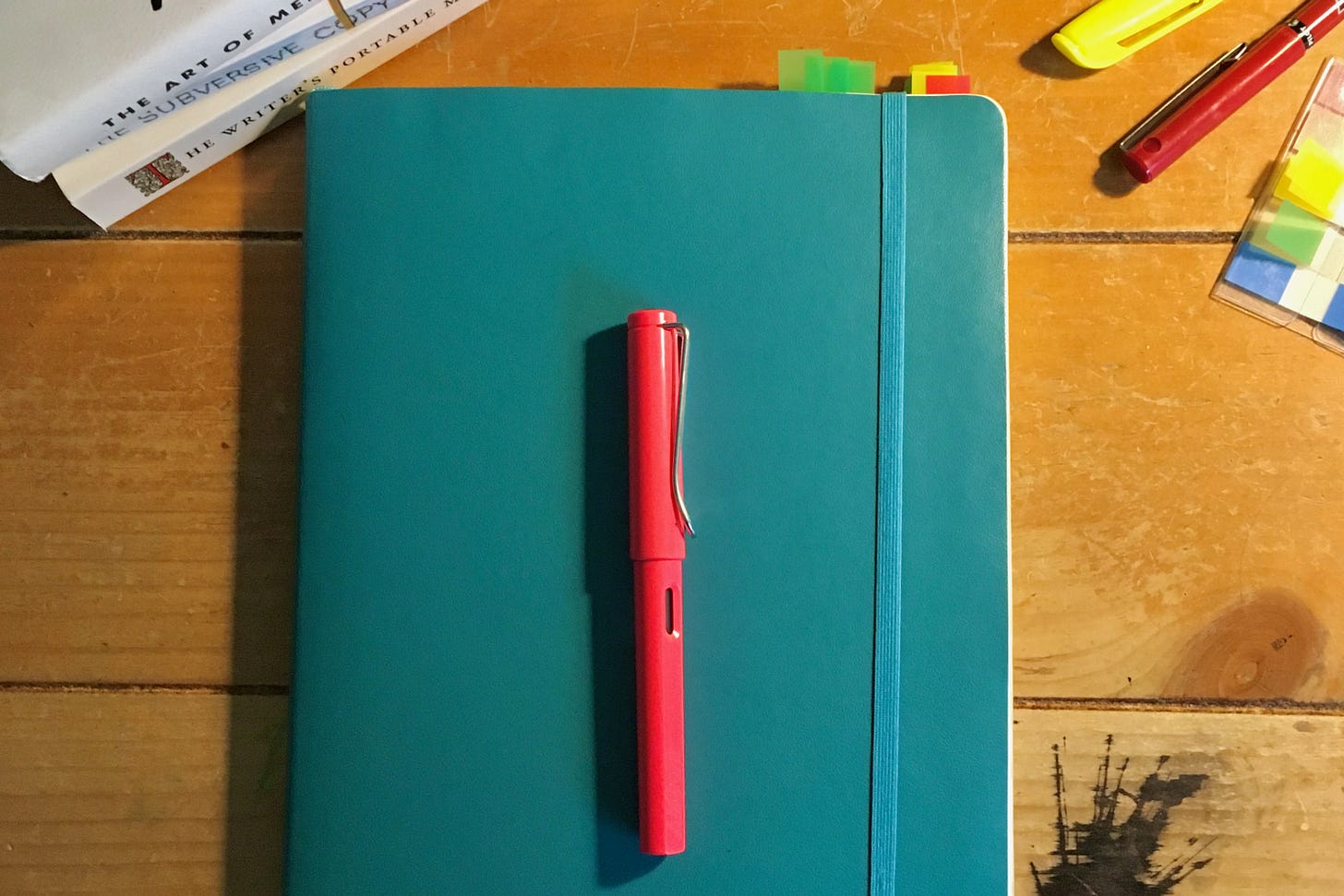 neon pink fountain pen on blue notebook surrounded by books, pens, and sticky tabs