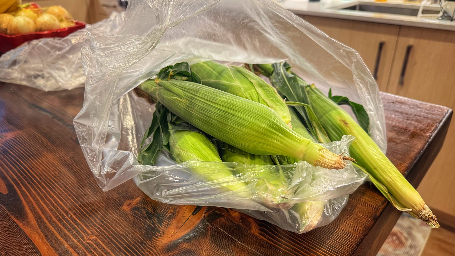 A plastic bag with several ears of unshucked corn is lying on a wooden kitchen table. A plastic bag with several ears of unshucked corn is lying on a wooden kitchen table.