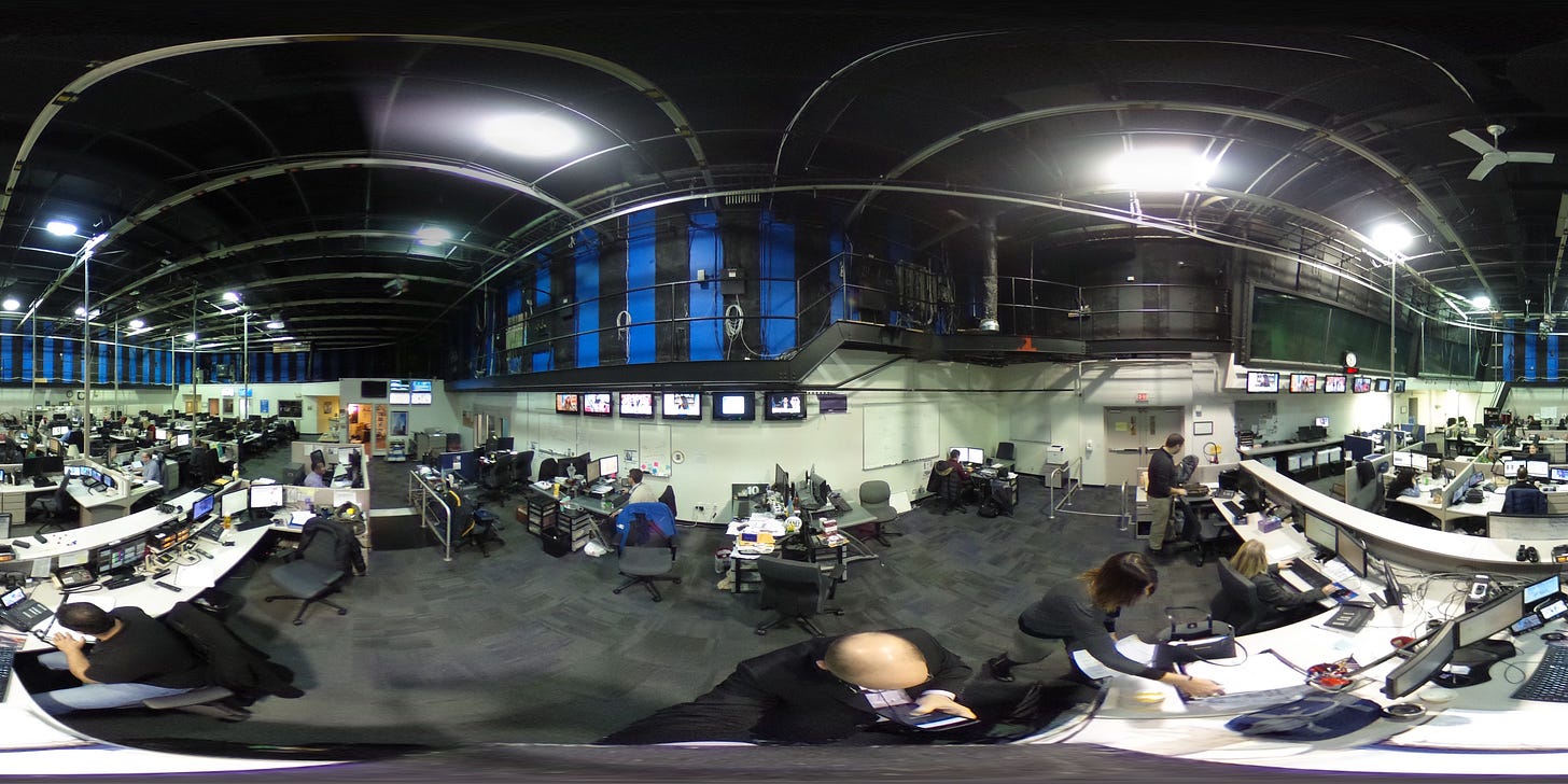 360-degree view of a busy television newsroom showing dozens of workstations with multiple monitors, people working at desks, TV screens mounted on walls, and industrial ceiling infrastructure in a large, open space with blue accent lighting. 360-degree view of a busy television newsroom showing dozens of workstations with multiple monitors, people working at desks, TV screens mounted on walls, and industrial ceiling infrastructure in a large, open space with blue accent lighting.