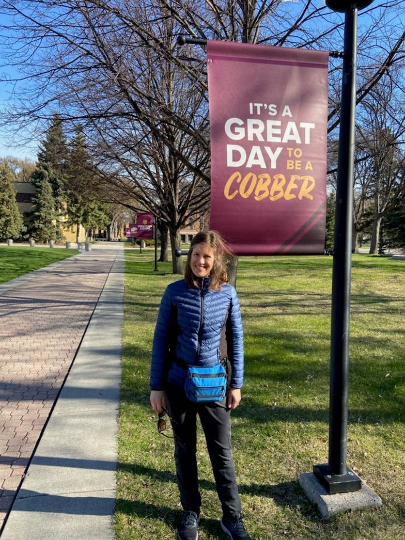 A woman (me) standing on a leafless college campus beside a sign that says "It's a great day to be a Cobber." A woman (me) standing on a leafless college campus beside a sign that says "It's a great day to be a Cobber."