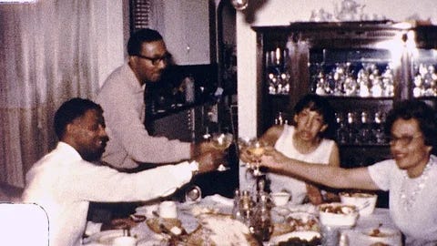 A grainy, warmly lit photo from what appears to be the 1970s shows four African American adults gathered around a dining table, raising their glasses in a toast during a holiday meal, likely Thanksgiving. A man on the left and a man in the center are smiling while clinking glasses with two women on the right side of the table. A roasted turkey is partially visible in the center foreground, and a dark wooden china cabinet filled with glassware stands in the background.