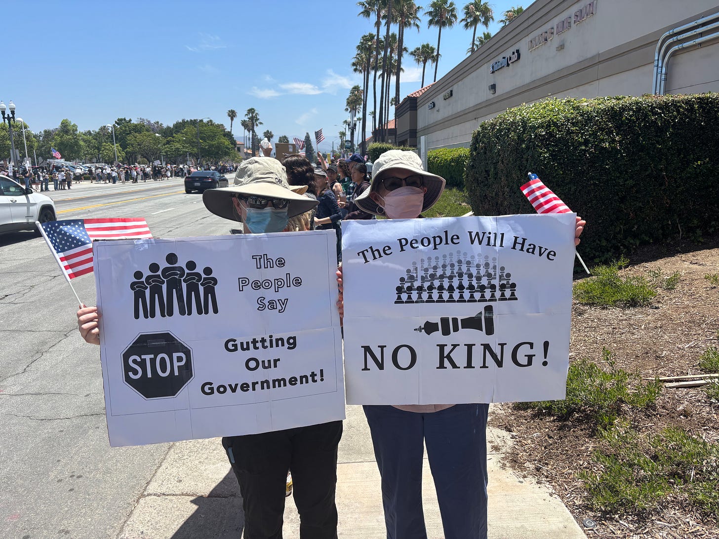 Two protesters in sun hats carrying American flags hold signs facing the camera. One reads "The People Say STOP GUTTING OUR GOVENRMENT." The other reads "The People Will Have NO KINGS," illustrated by a fallen king chess piece in front of an army of other chess pieces. Behind them, protesters also carrying American flags line both sides of a palm-tree lined street. Two protesters in sun hats carrying American flags hold signs facing the camera. One reads "The People Say STOP GUTTING OUR GOVENRMENT." The other reads "The People Will Have NO KINGS," illustrated by a fallen king chess piece in front of an army of other chess pieces. Behind them, protesters also carrying American flags line both sides of a palm-tree lined street.