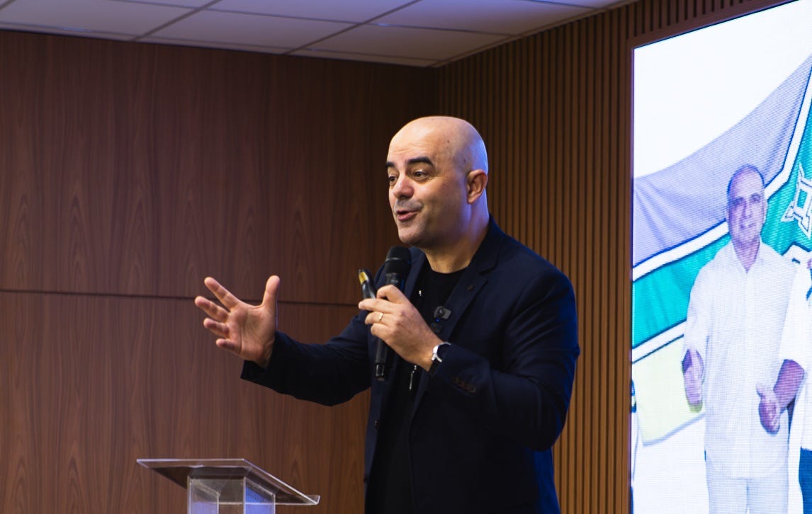 Palestrante de Vendas Leandro Branquinho falando de forma animada com a mão estendida e um microfone na outra mão