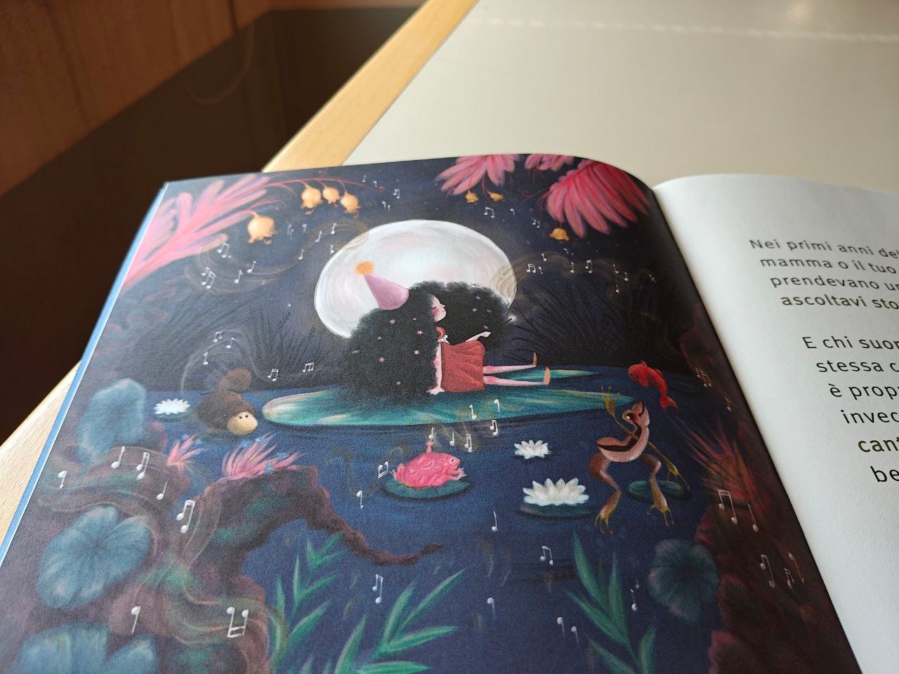 Pagina illustrata della Scatolina NOTA edita da effequ, appoggiata su un tavolo di legno color bianco sporco, con un pezzetto di muro in ombra nell'angolo in alto a sinistra. Nel disegno compare uno stagno blu con una grande ninfea al centro, su cui è seduta una bambina dalla pelle chiara con una foltissima chioma di capelli neri che casca fino a terra, e porta un cappellino rosa con un pompon arancio. Attorno a lei volteggiano nell'aria note musicali bianche e ondeggiano piante marroni e rosa nei quattro angoli, alcune con campanule gialle alle estremità. Nello stagno vediamo una rana che salta, un pesce rosso che esce dall'acqua e un animaletto con la corazza marrone e il volto giallo. Sullo sfondo notturno campeggia l'enorme disco giallo pallido della luna. Pagina illustrata della Scatolina NOTA edita da effequ, appoggiata su un tavolo di legno color bianco sporco, con un pezzetto di muro in ombra nell'angolo in alto a sinistra. Nel disegno compare uno stagno blu con una grande ninfea al centro, su cui è seduta una bambina dalla pelle chiara con una foltissima chioma di capelli neri che casca fino a terra, e porta un cappellino rosa con un pompon arancio. Attorno a lei volteggiano nell'aria note musicali bianche e ondeggiano piante marroni e rosa nei quattro angoli, alcune con campanule gialle alle estremità. Nello stagno vediamo una rana che salta, un pesce rosso che esce dall'acqua e un animaletto con la corazza marrone e il volto giallo. Sullo sfondo notturno campeggia l'enorme disco giallo pallido della luna.