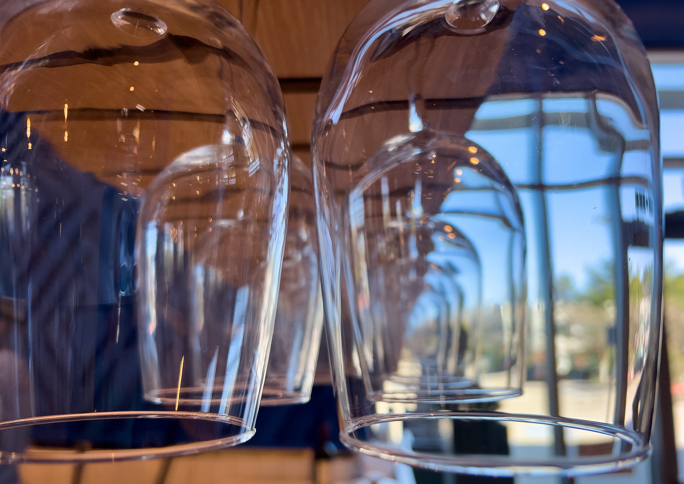 Several upside-down wine glasses hang in a row with a blurred background of windows and outdoor scenery.