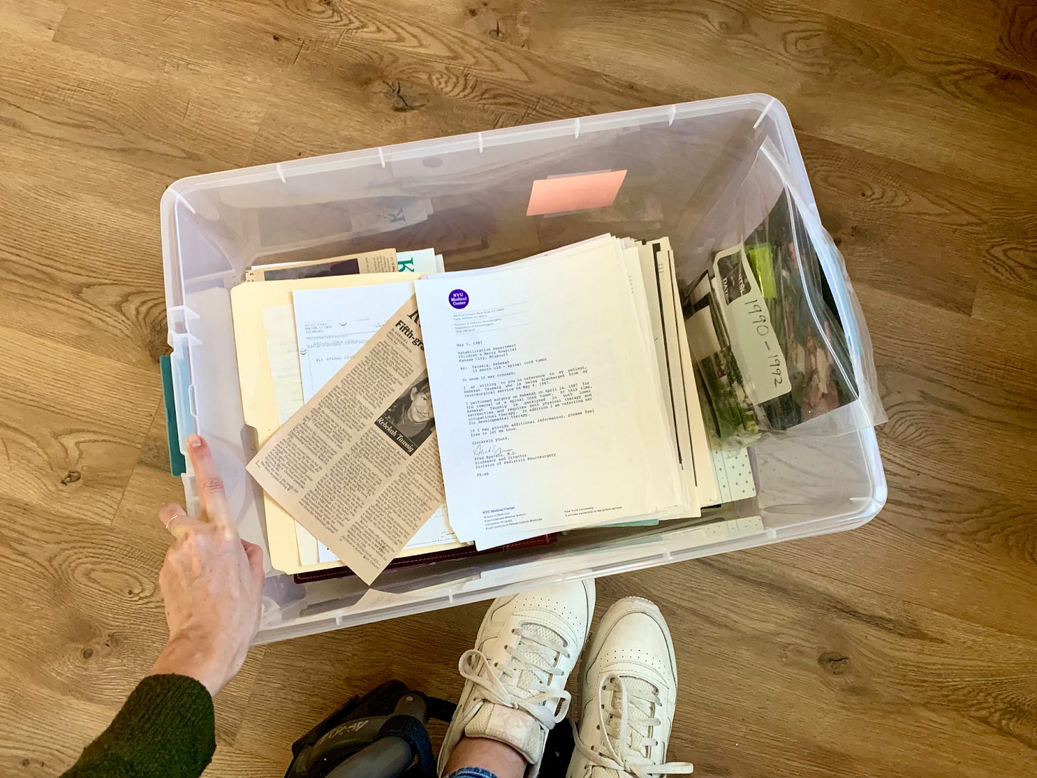 A photo taken from above of the tub of old artifacts her mom gave her. A newspaper clipping with a photo of Rebekah in 5th grade peeps through medical documents. A bag of photos labeled 1990-1992 rests on the right side of the tub. Rebekah's left hand rests on the side of the tub, the tops of her white tennis shoes and wheelchair frame peek into the bottom of the photo. A photo taken from above of the tub of old artifacts her mom gave her. A newspaper clipping with a photo of Rebekah in 5th grade peeps through medical documents. A bag of photos labeled 1990-1992 rests on the right side of the tub. Rebekah's left hand rests on the side of the tub, the tops of her white tennis shoes and wheelchair frame peek into the bottom of the photo.