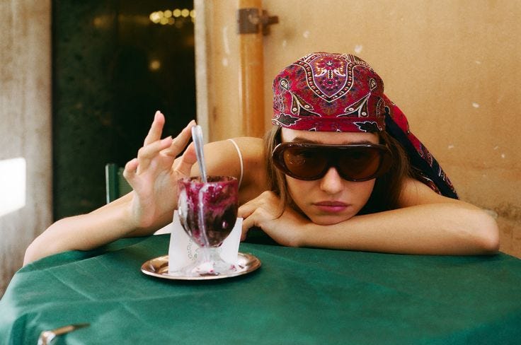 This may contain: a woman wearing sunglasses and a bandana leaning on a table with a drink in her hand