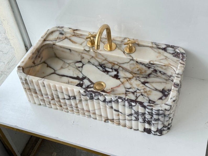 May include: A white marble sink with a gold faucet and drain. The sink has a fluted edge and is rectangular in shape.