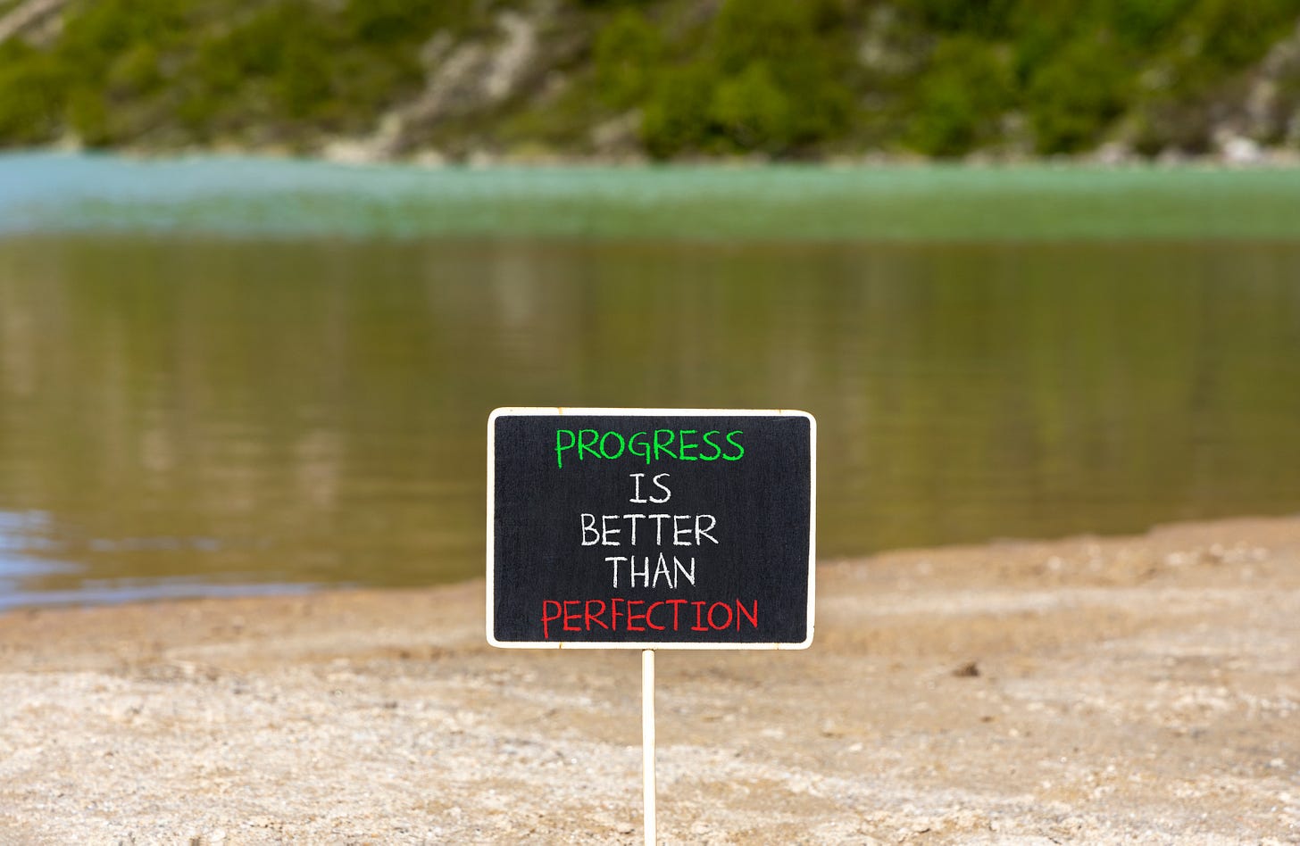 sign reading Progress is better than Perfection on a beach with a water background
