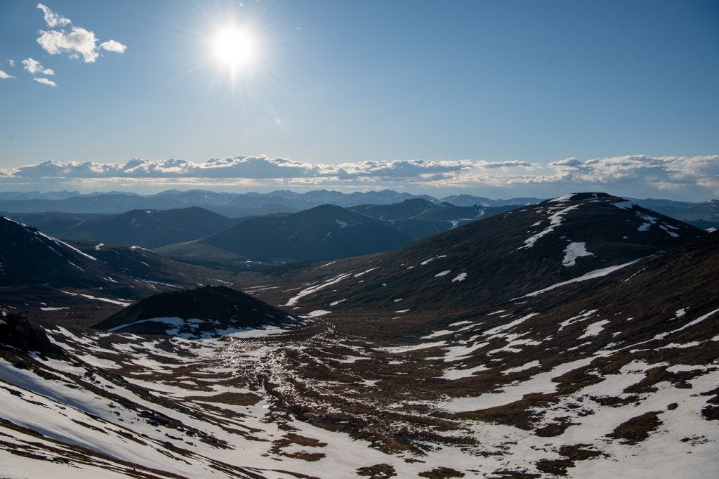 The low sun stars brightly above a long valley veined with snow and braided meltwater, rows of dark ridges layering back to the horizon.