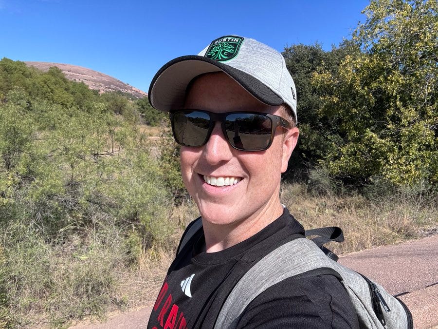 A man in his mid-40s with short hair and a smiling expression wears dark sunglasses, a gray cap featuring a green circular patch with white text and emblem, and a black short-sleeve shirt. He stands on a reddish dirt path in a natural outdoor setting with green bushes, trees, rocky hills, and distant mountains under a clear blue sky. A gray backpack with straps is visible on his back.