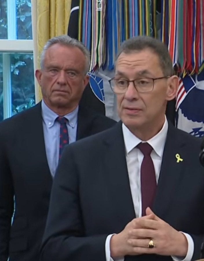 Robert Kennedy and the CEO of Pfizer standing side by side. Both men wear suits, ties, and glasses. A backdrop of colorful ribbons and flags is visible behind them.