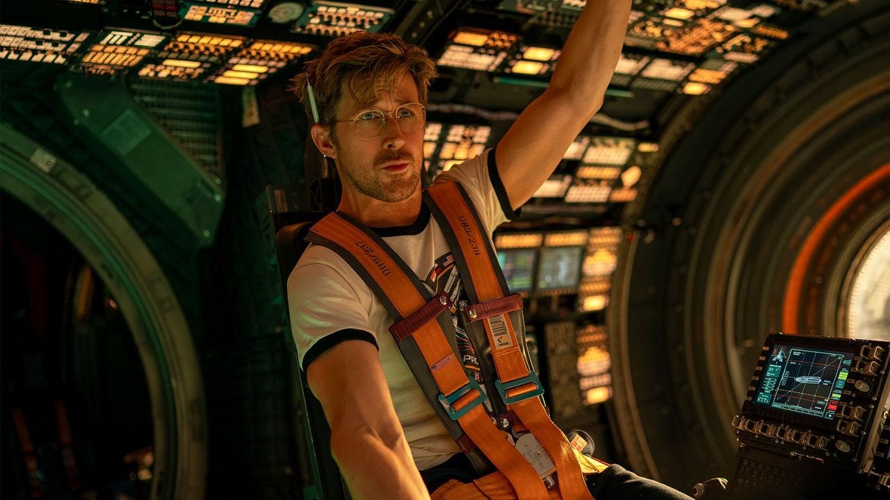 Gosling in glasses, short sleeves, orange safety thing, looking intense sitting in spaceship interior