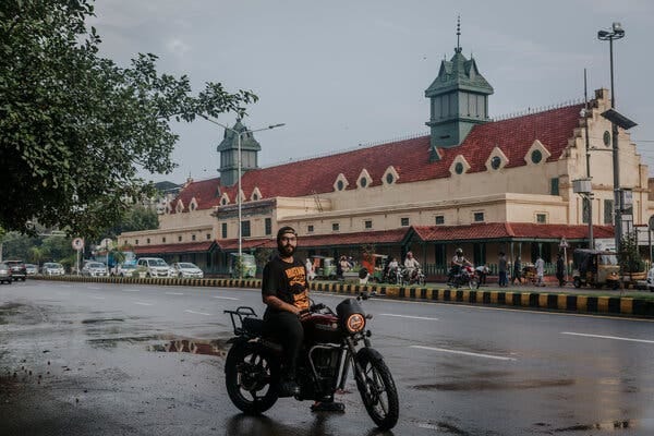 Portrait of a man astride a motorcycle in front of a colonial-era building with tile roof.