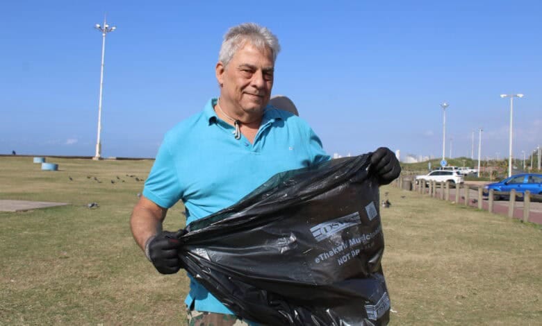 A naturist man in a teal shirt and gloves holds a trash bag while participating in a community cleanup near Umhlanga Lagoon on a sunny day. A naturist man in a teal shirt and gloves holds a trash bag while participating in a community cleanup near Umhlanga Lagoon on a sunny day.
