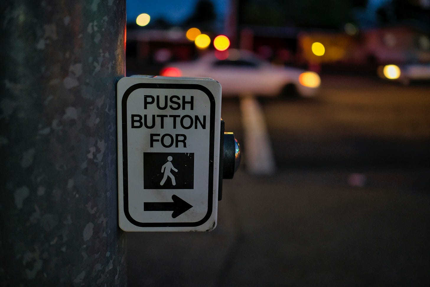 Sfondo sfuocato di un ambiente urbano notturno con strada, una macchina bianca e dei punti luce. In primo piano, sulla sinistra, un palo con un bottone da premere e un cartello con scritto PUSH BUTTON FOR e il disegno di un omino che attraversa. Sfondo sfuocato di un ambiente urbano notturno con strada, una macchina bianca e dei punti luce. In primo piano, sulla sinistra, un palo con un bottone da premere e un cartello con scritto PUSH BUTTON FOR e il disegno di un omino che attraversa.