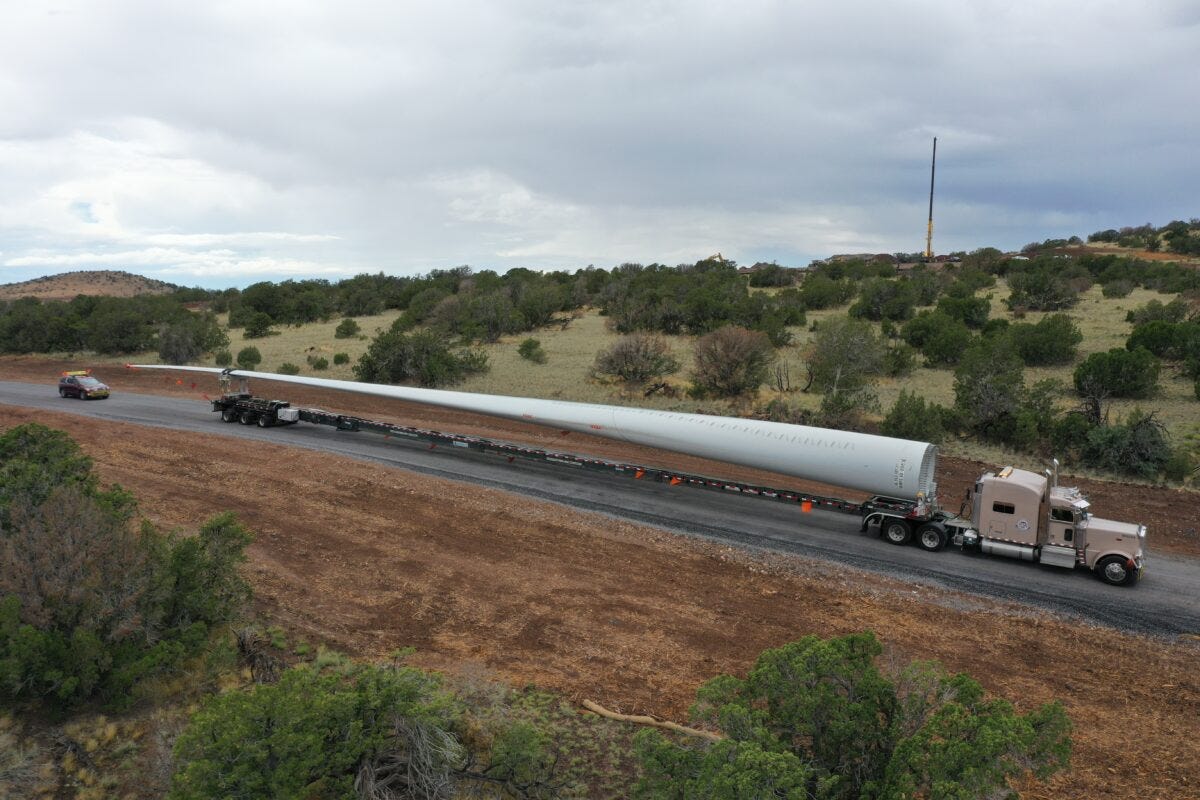 A semi-tuck hauls a long, white, narrow wind turbine blade, with a small car behind it. The wind turbine blade is probably 20 times the length of the car.