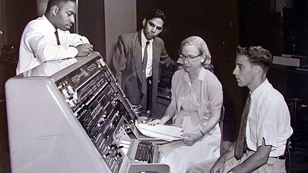 Black and white photo of computer programmer and co-creator of the COBOL programming language Grace Hopper. She is a white woman who is seated at an old fashioned computer terminal with three young men (one Black, one South Asian, one white) who all look like they are taking instruction from her.
