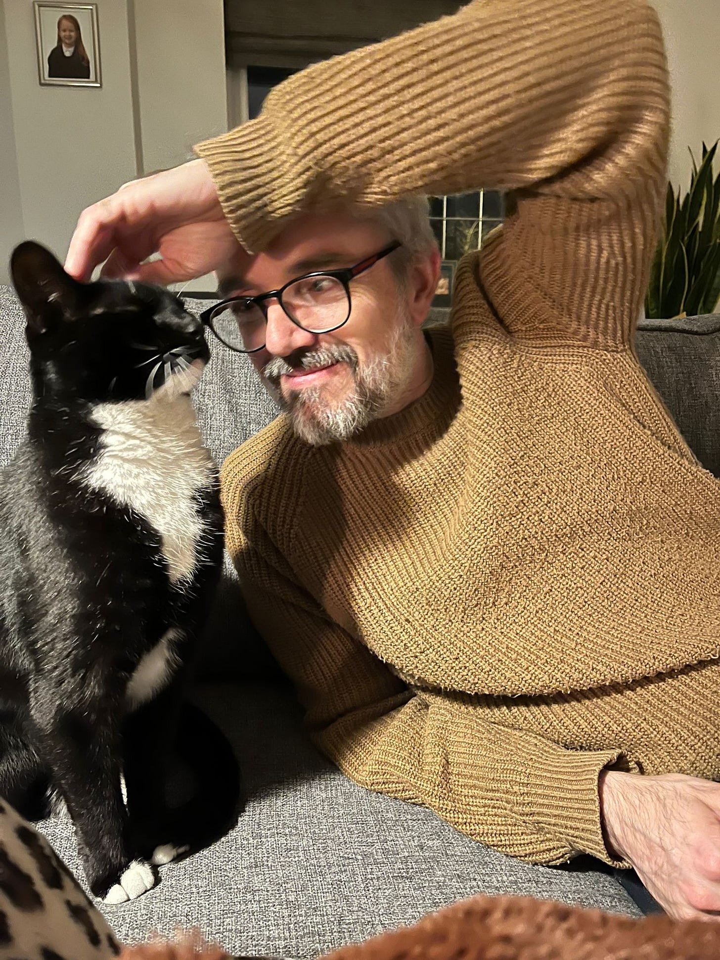 A bearded middle aged man with glasses, grey hair and a beard, tickling the head of a tuxedo cat. The man is the author, Ollie, and the cat is called Henry. They are sitting on a sofa.