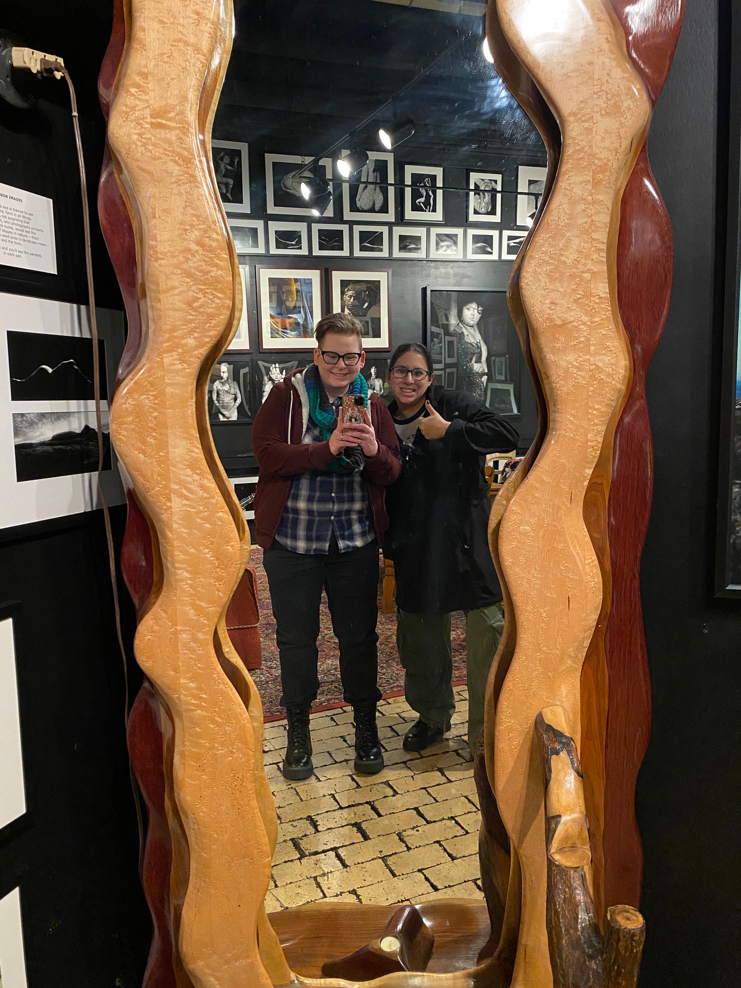Photo of two people reflected in a mirror, one giving a thumbs up. Behind are black and white photos on the wall in the Marilyn Monroe Room of Artisan Works in Rochester, NY.