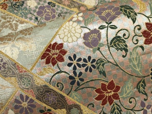 Close-up of a Saga Nishiki six-panel obi: diamond-framed compartments each containing a distinct floral composition — chrysanthemum, peony, clematis — woven in polychrome silk and metallic thread against alternating pink and silver-green grounds. The panel divisions themselves, bordered in gold, demonstrate the architectural logic of classical motif arrangement. Close-up of a Saga Nishiki six-panel obi: diamond-framed compartments each containing a distinct floral composition — chrysanthemum, peony, clematis — woven in polychrome silk and metallic thread against alternating pink and silver-green grounds. The panel divisions themselves, bordered in gold, demonstrate the architectural logic of classical motif arrangement.
