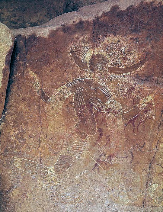 Dark red and white painting on a brown stone wall. The figure is a dark red woman wearing white beads and fringe, with dark red horns coming out of her head and white dots raining down above her.
