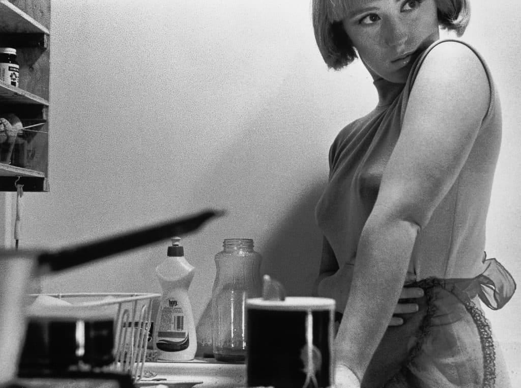Foto in bianco e nero di una ragazza in cucina, raffigurata dalla fronte a sopra in ginocchio.  Si trova davanti al lavandino, si vede il detersivo e qualche utensile. Ha la frangetta e un caschetto, capelli lisci e chiari. Un velo di trucco. Un vestito chiaro, smanicato, con un grembiule.