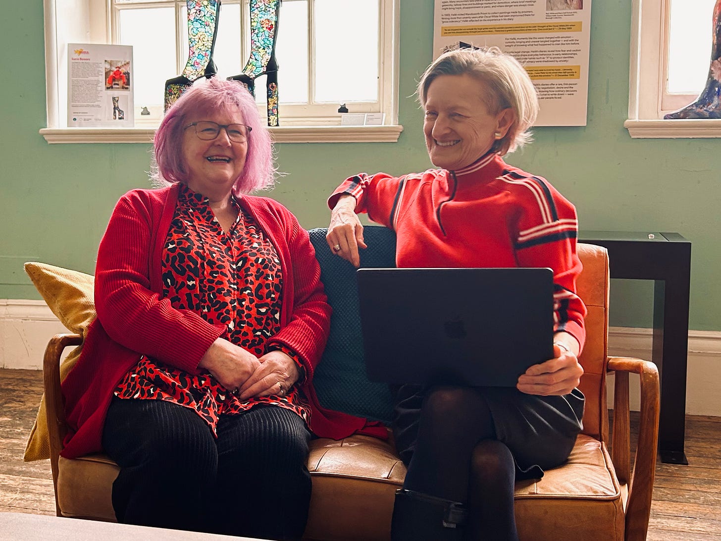 Two women, the author and Jeanne, sitting together on a tan leather sofa and laughing. Both are wearing red tops; Jeanne is in a leopard-print blouse and cardigan, and the author is in a red zip-up athletic jacket, holding a laptop.