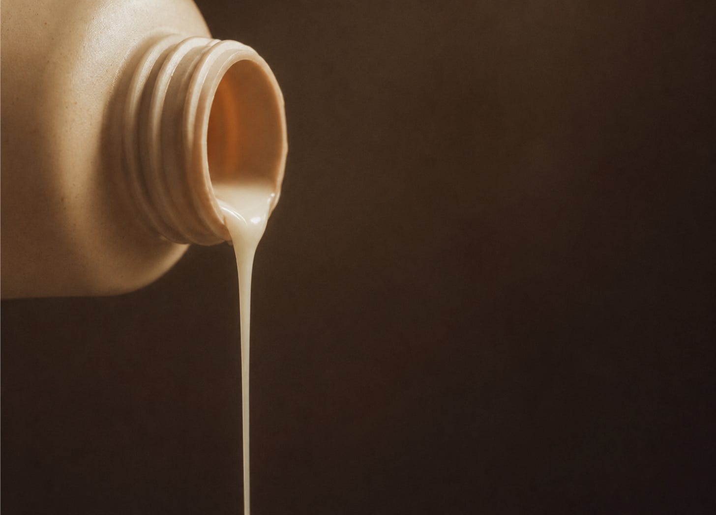 Minimalist photograph of a tan bottle tipped on its side, with creamy liquid pouring out against a soft dark background. Minimalist photograph of a tan bottle tipped on its side, with creamy liquid pouring out against a soft dark background.