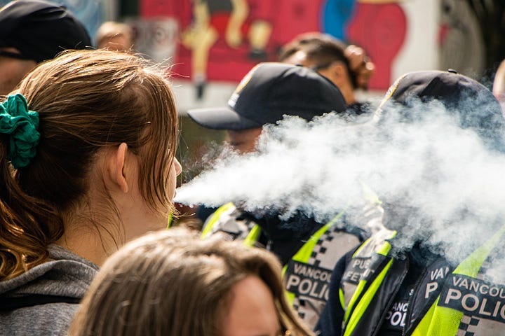 First image: A woman blows a cloud of vapour in the faces of police. A friend who was down there tells me her group was trolling the police the whole time, including taking drags from their vape and blowing it at the police. Second image: A person with a white shirt and a white wrap around their head and face and a black cap holds up a sign that reads: "Cops are better at committing crimes than solving them." Third image: An Indigenous person, back facing the line of police, is holding a bowl of smoking sage. After holding a cap overtop of the sage smoke, they turned the cap over, and smoke can be seen trickling upwards, out of the hat. Fourth image: A couple of police officers are seen at the front line. One is wearing a mask and looking dead ahead. The other is looking at something to the left of the camera's perspective.