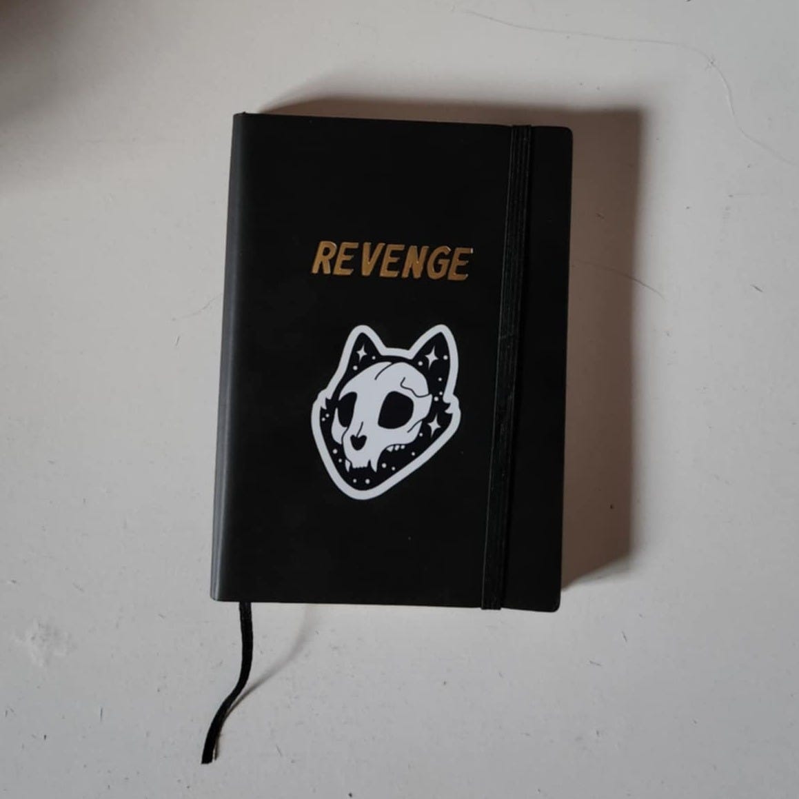 A black notebook with the word "Revenge" written in gold script and a cat skull sticker underneath.