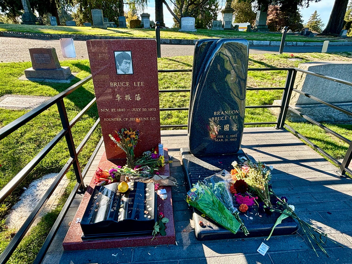 color photo of two grave markers for Bruce Lee and Brandon Lee, with flowers, coins, and other things left as offerings