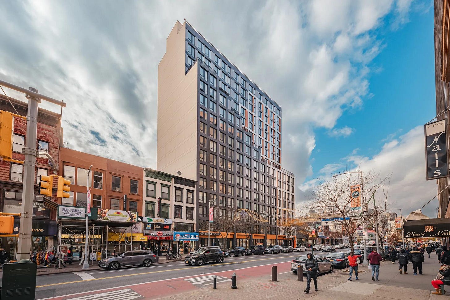 Street view of a mixed-use block in Harlem, with pedestrians on a wide sidewalk, storefronts at ground level, cars along the street, and a large apartment building rising above older row buildings.