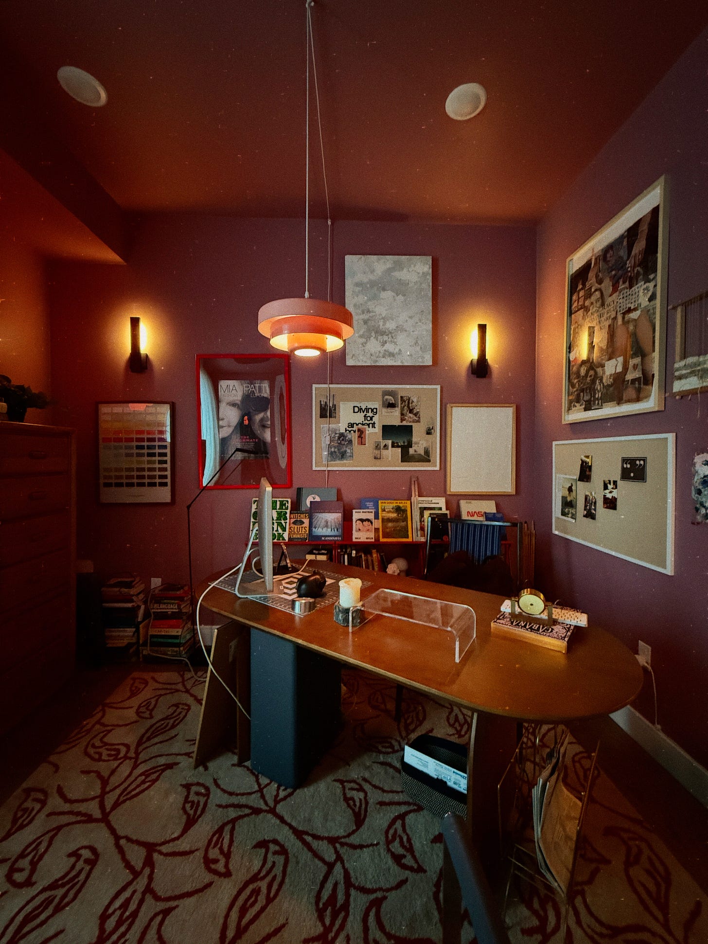 Melissa Geurts' warmly lit home office with burgundy walls and wooden desk, featuring an extensive collection of framed artwork covering the walls including gallery posters, personal art collages, paintings from various artists, and pieces from past discoveries, all illuminated by an orange pendant light Melissa Geurts' warmly lit home office with burgundy walls and wooden desk, featuring an extensive collection of framed artwork covering the walls including gallery posters, personal art collages, paintings from various artists, and pieces from past discoveries, all illuminated by an orange pendant light