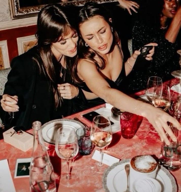 This may contain: two young women sitting at a table with wine glasses and plates on it, posing for the camera