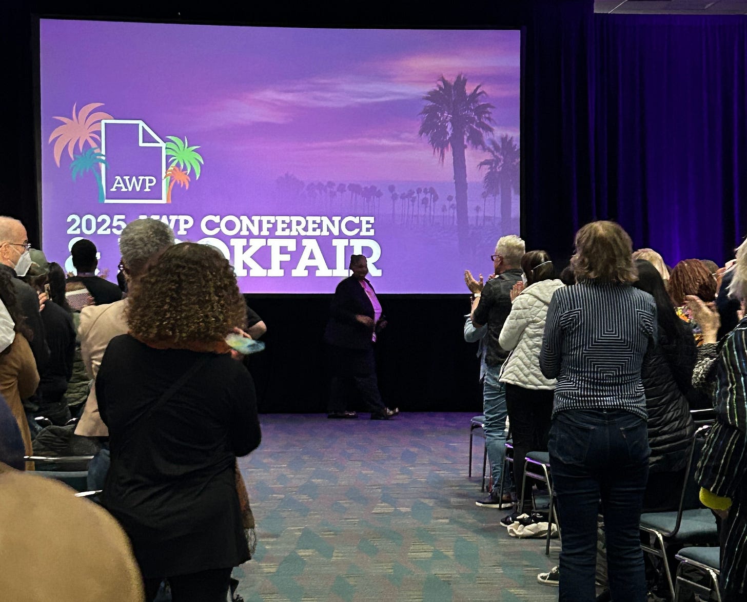 Roxane Gay walking off the stage with a purple AWP background behind her and audience members standing and applauding.